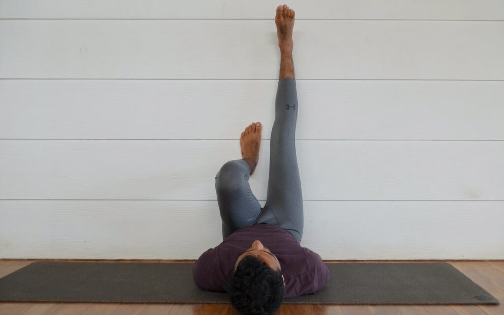 Man in gray leggings and a maroon shirt practicing a modification and variation for Legs Up The Wall Pose with his left leg bent in Blue Osa's yoga shala in Costa Rica.