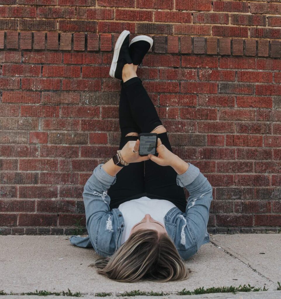 Woman with a jean jacket and black jeans with her legs up a brick wall while looking at her cell phone showing that you can practice Legs Up The Wall any time, any where.