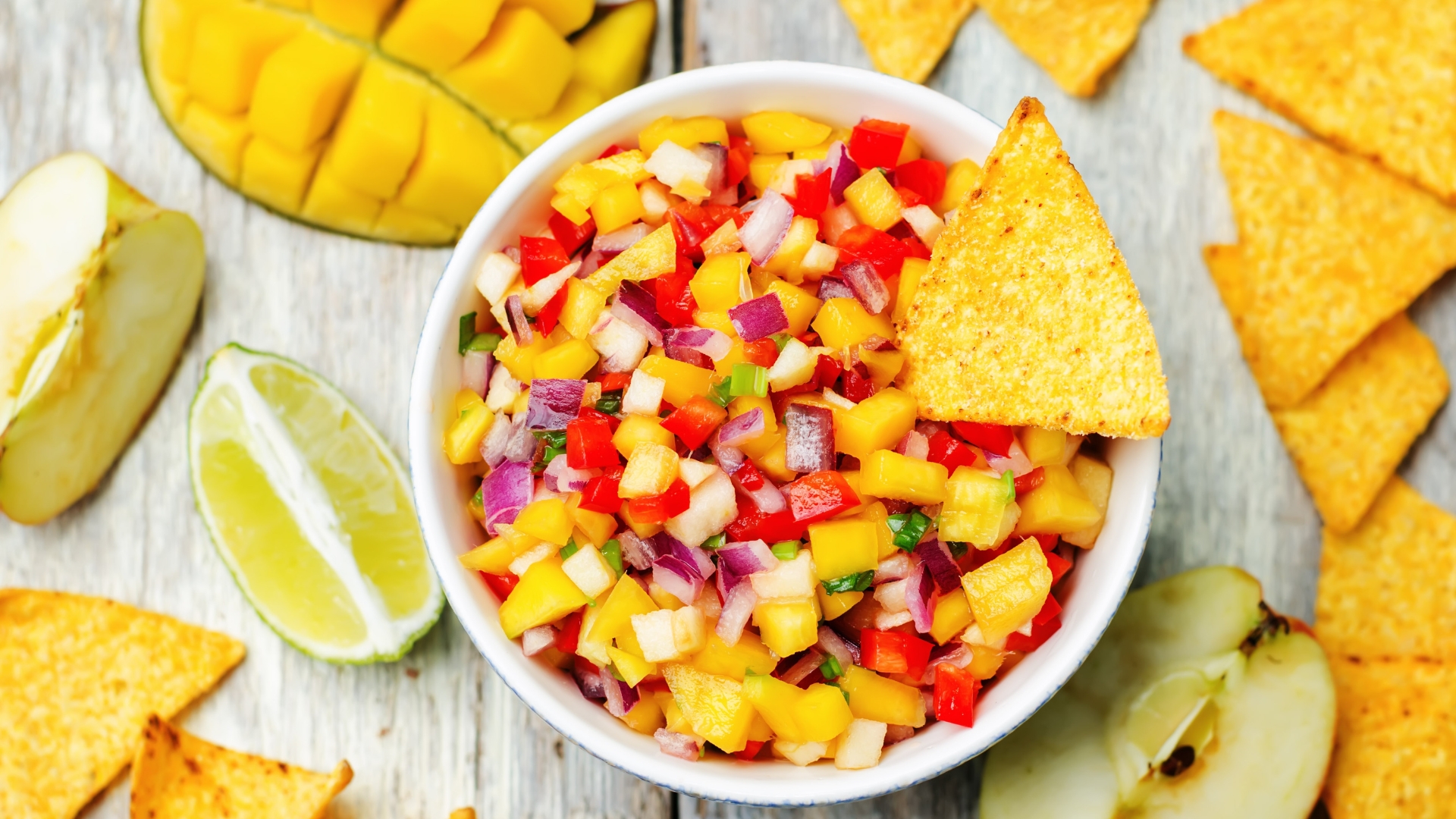White ceramic bowl of mango salsa surrounded by yellow tortilla chips and a sliced mango.
