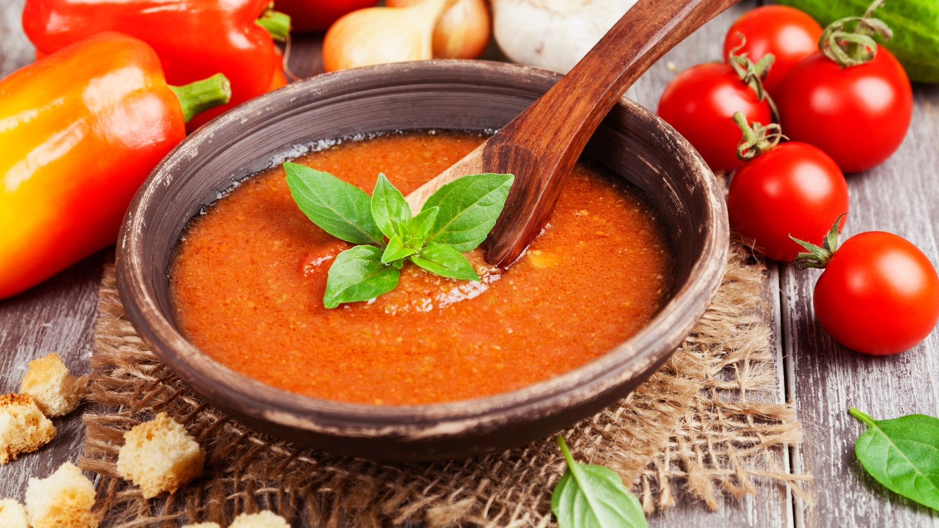 Brown ceramic bowl of reddish orange gazpacho soup with tomatoes and peppers surrounding.
