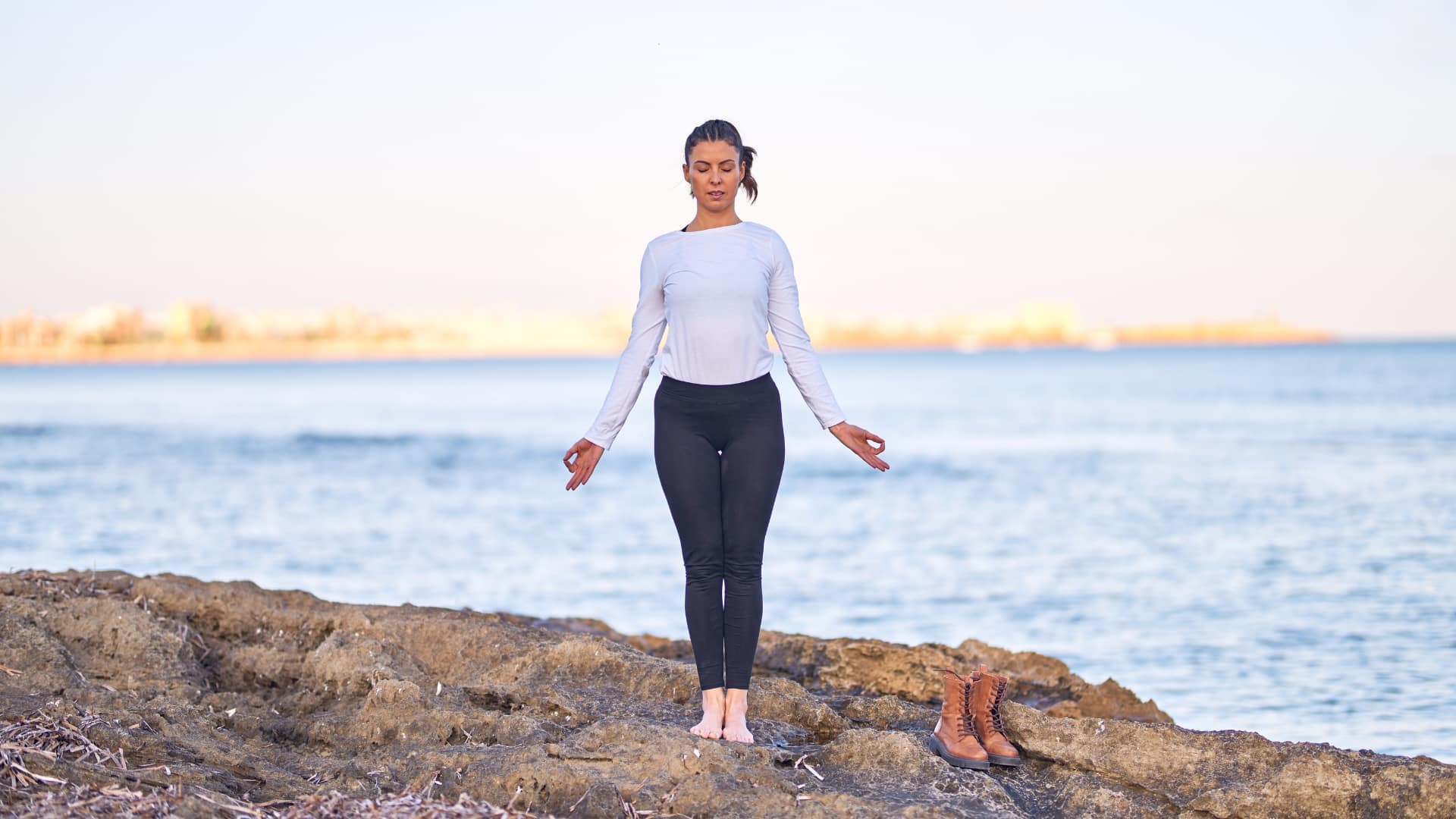 Woman in a white long sleeve tee and black leggings standing in mountain pose on a rocky surface in front of a body of water.