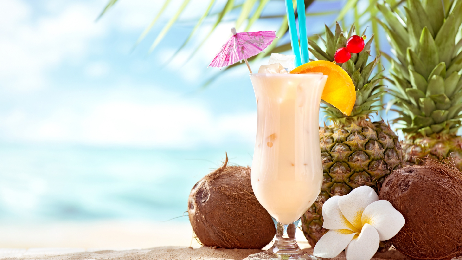 Pina Colada on a beach with a white tropical flower, coconuts and a pineapple sitting next to it.