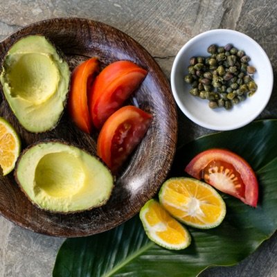 Wooden bowl filled with sliced avocado, tomatoes and lemon. A small white bowl of capers and sliced tomato and lemon on the side. 