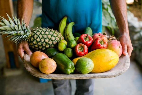Torso of a man in a blue shirt holding a large wooden plate of fruit and vegetables.
