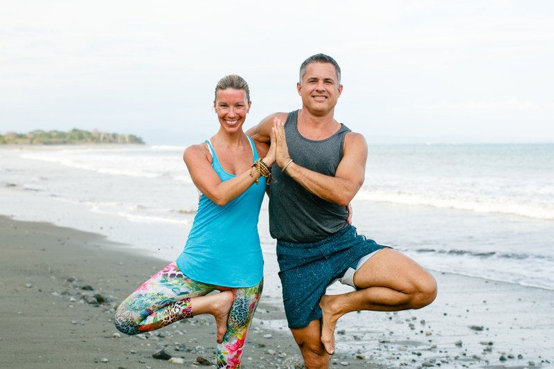 A man and a woman standing side by side in tree pose touching hands on the beach in Costa Rica. 