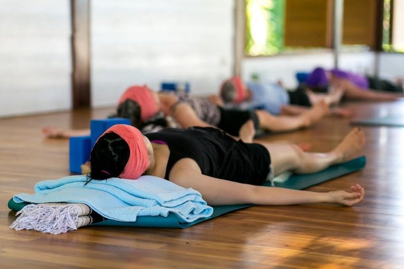 Woman laying in savasana with a pink towel wrapped around her eyes and a blue blanket under her head in Blue Osa's yoga shala in Costa Rica. 