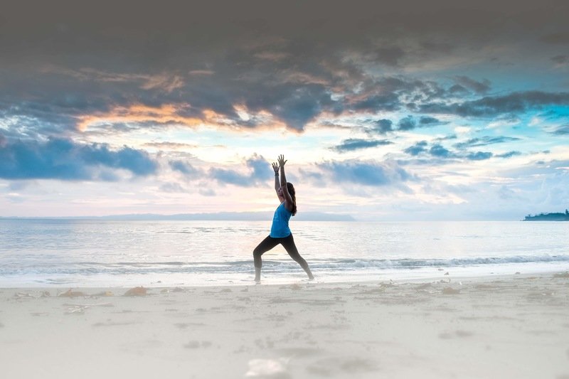 Woman in a blue tank top and black leggings practicing warrior one on the beach at sunset in Costa Rica. 