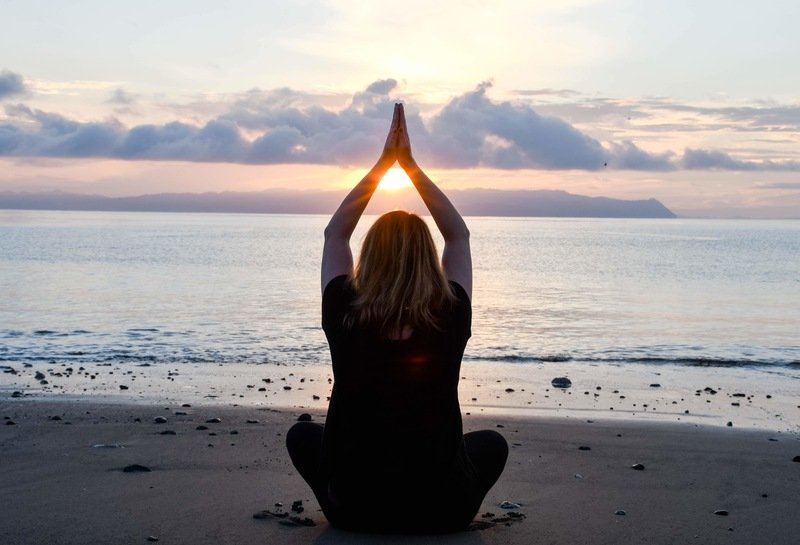 Woman sitting in a cross legged meditation pose on the beach with her hands above her head in prayer position with the setting sun shining through her arms in Costa Rica. 