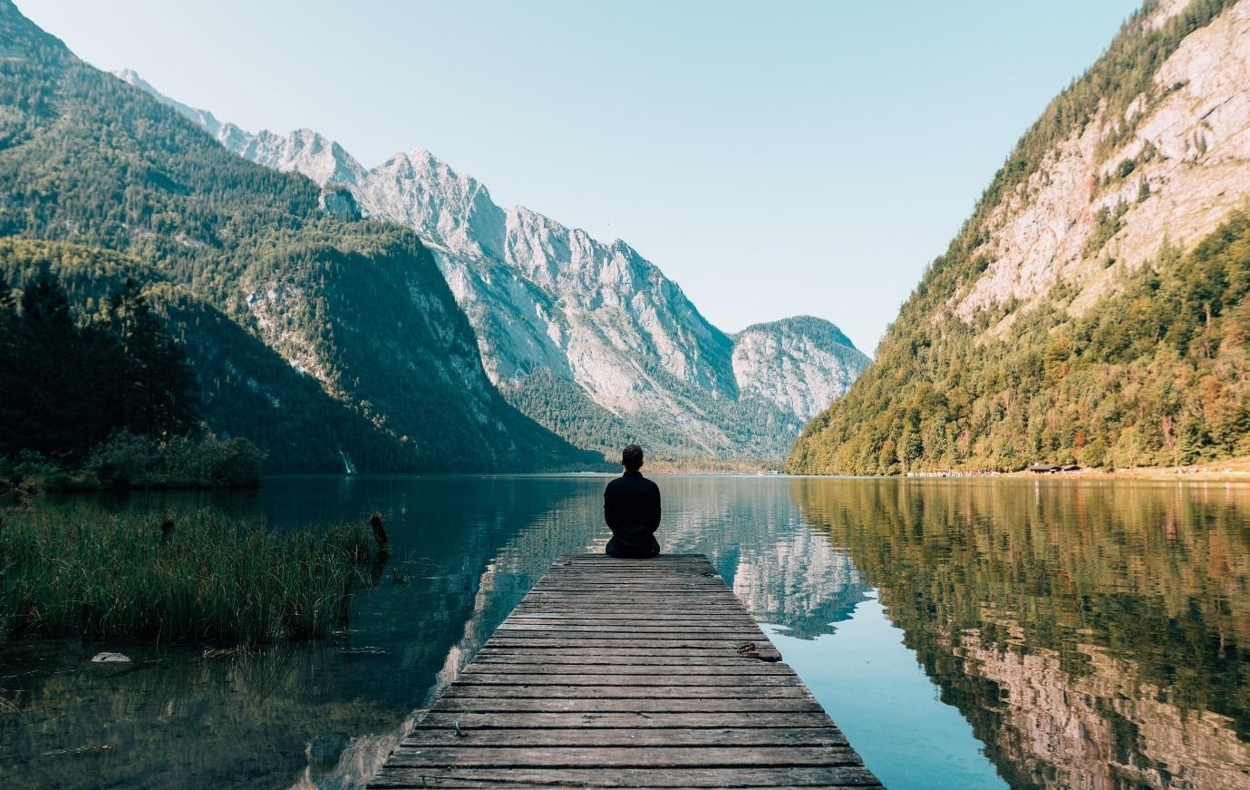 Man sitting on the edge of a dock overlooking a river and mountains.