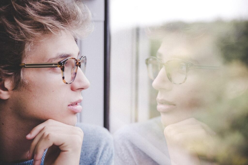 Young man with glasses looking out a window with his reflection staring back at him. 