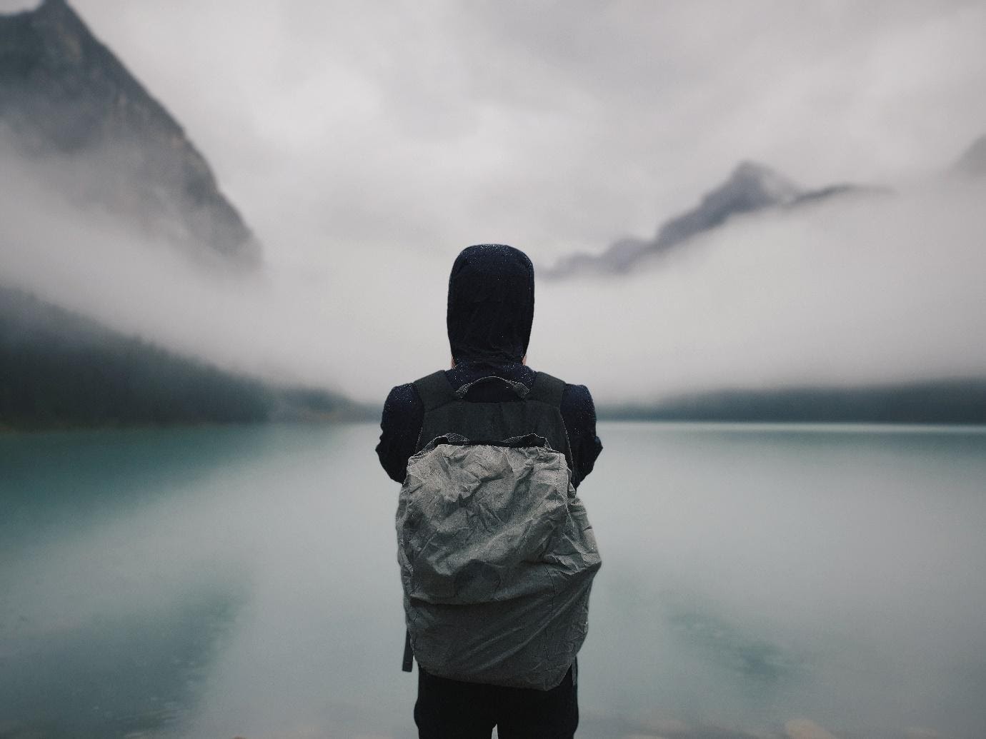 A person standing in a black hoodie with a backpack on staring out into a foggy nature environment