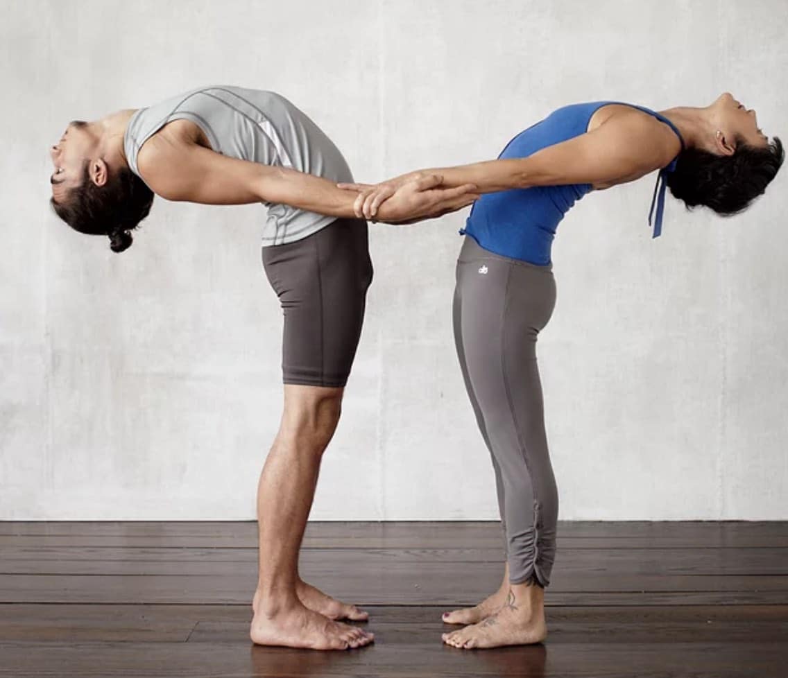 Two people are shown face to face while doing a series of partner yoga poses. The partners are holding each other's forearms while the front partner is performing a backbend