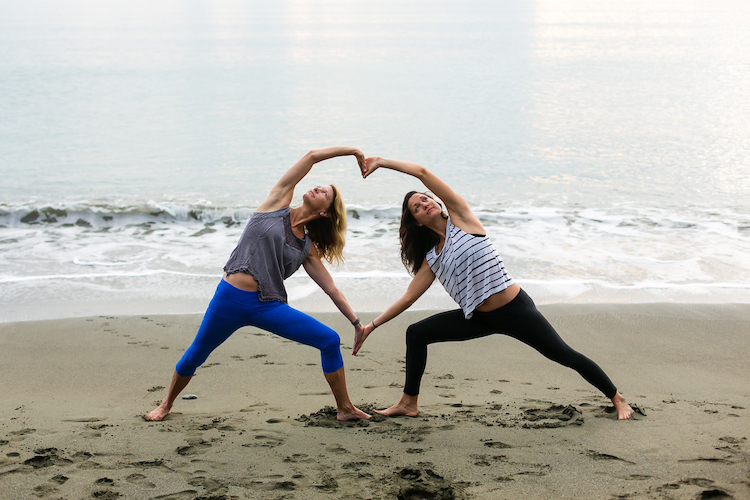 Two women standing in a side angle lunge with their arms together making a heart shape on the beach in Costa Rica. 
