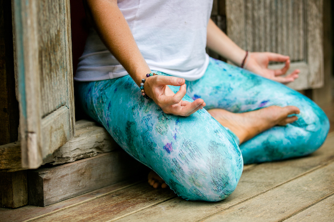 The lower half of a woman's body wearing turquoise leggings sitting in a meditative position and hands in a thumb to pointer finger mudra. 
