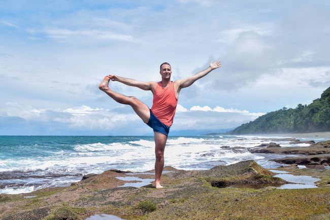 Yogi Aaron practicing extending hand to big toe pose on a beach in Costa Rica. 