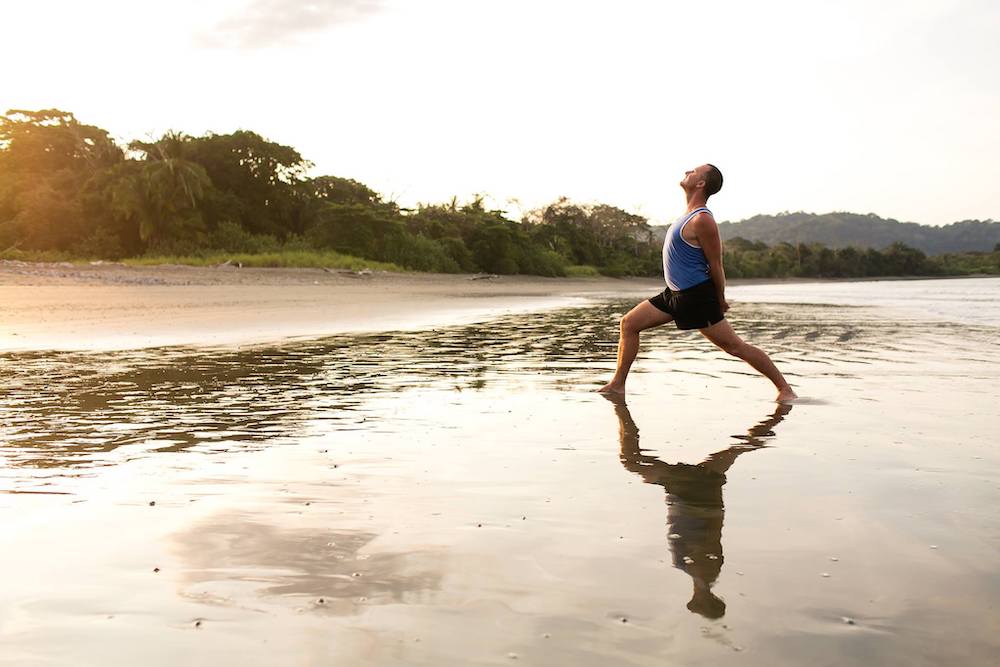 Yogi Aaron standing in a warrior one  variation pose with his hands behind his back standing in the water on a beach in Costa Rica. 