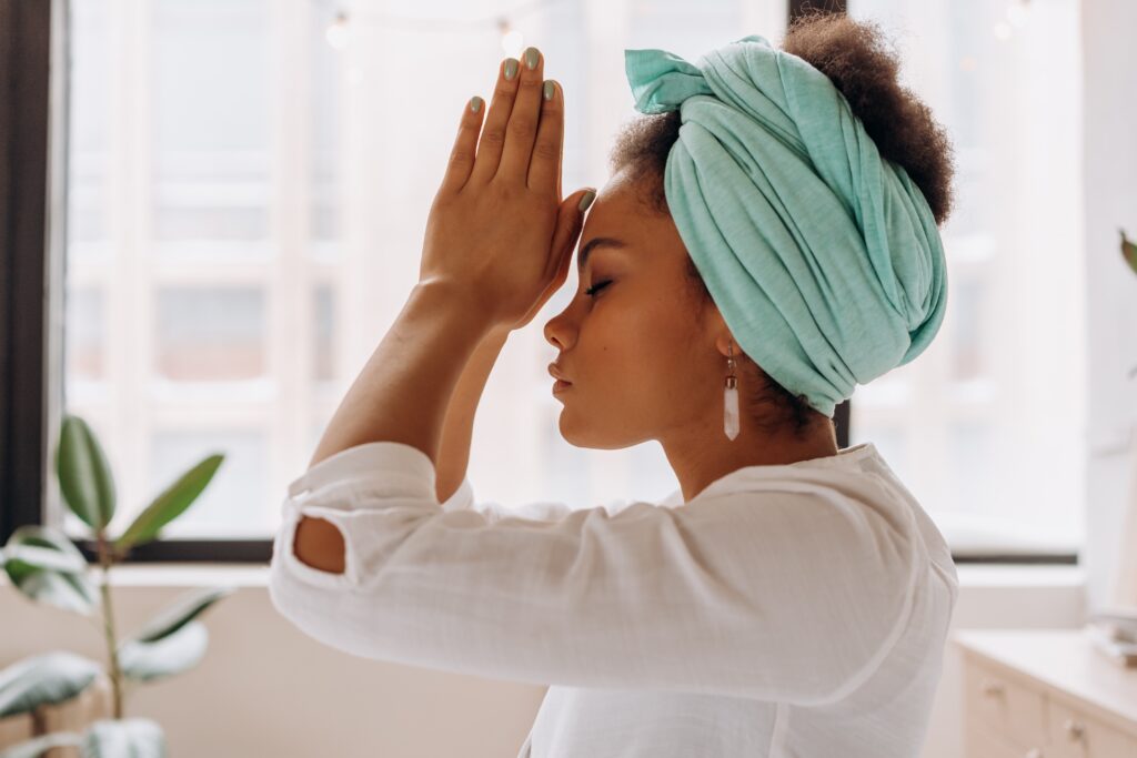 Woman wearing a turquoise head wrap and a white shirt with her hands in prayer position at her third eye. 