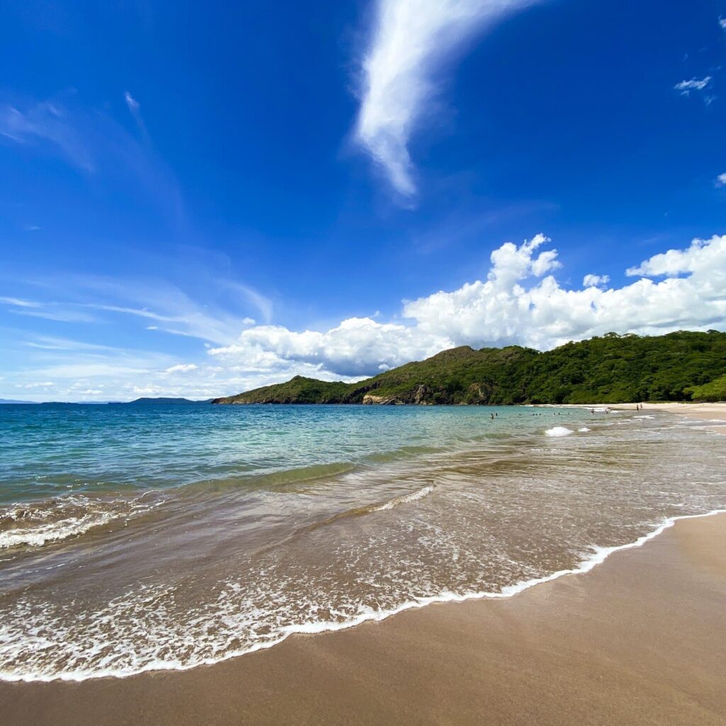 Pristine beach in Costa Rica with turquoise water washing over tan sand with lush green mountains in the distance.