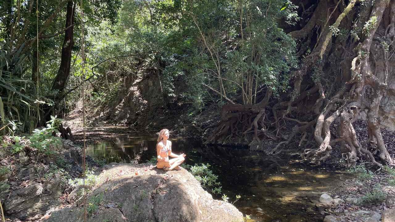 Woman meditating in a cross legged position on a rock next to a river and giant tree in Costa Rica.