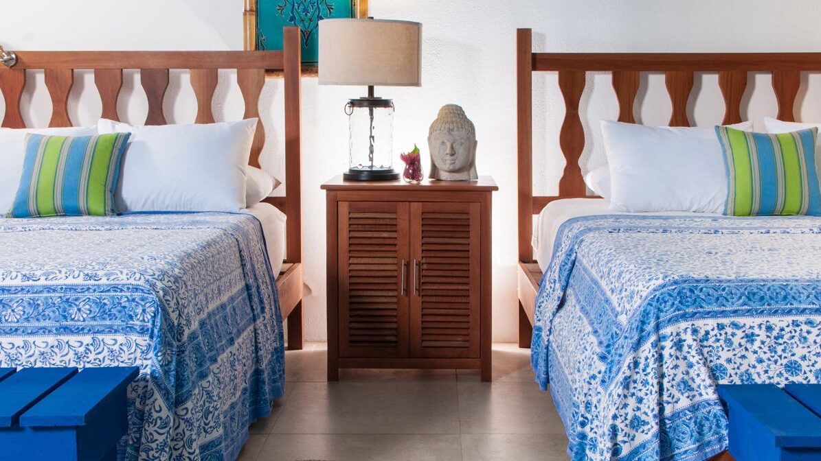 A room at Blue Osa Yoga Retreat Center in Costa Rica with two queen beds, blue and green bedding and wood furniture with a buddah head and lamp on the nightstand. 