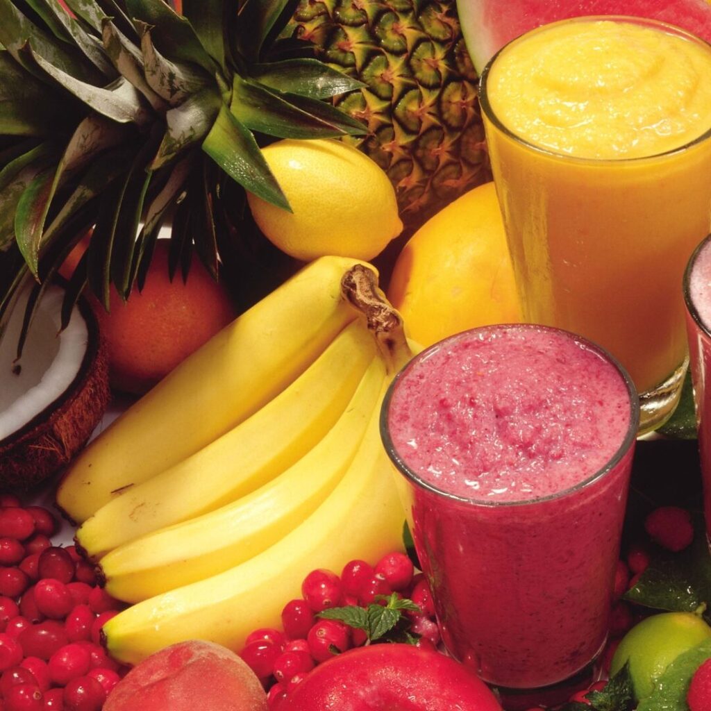 Bananas, lemon, pineapples and berries next to two glasses, one of a pink smoothie and one of a yellow smoothie.