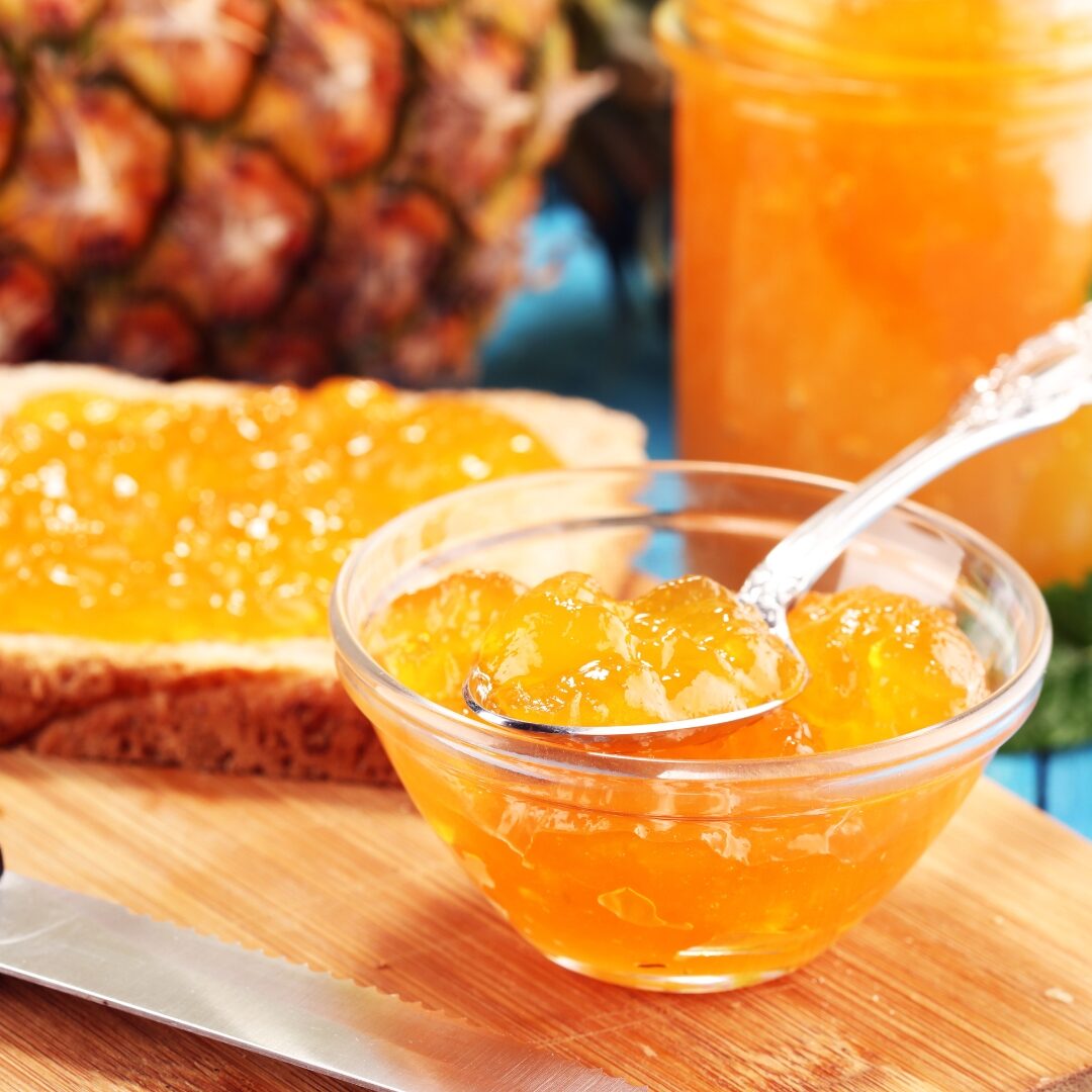Glass dish of pineapple jam with a slice of bread with pineapple jam and a pineapple in the background. 