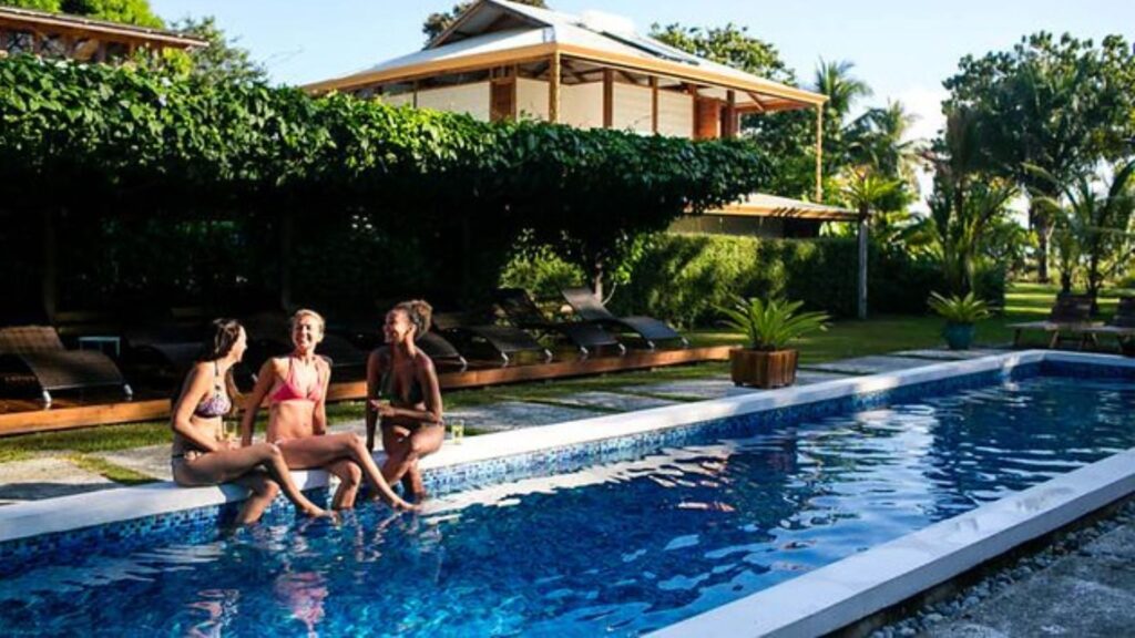 Three women sitting poolside together and laughing with their legs in the water of Blue Osa's chemical free lap pool. 