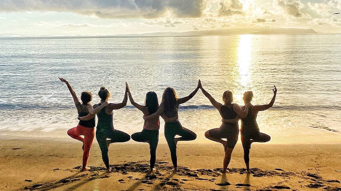 Six yoga students practicing tree pose connected by their hands looking out into the sunset on the beach at Blue Osa Yoga Retreat Center in Costa Rica. 