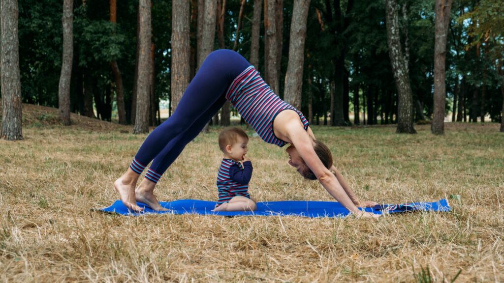 A mom doing a down dog yoga pose over top of her baby in a field. 