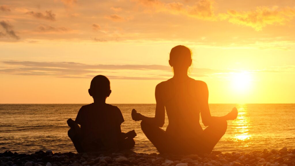 A mom and her child sitting side by side in a cross legged meditation pose over looking a sunset over the ocean. 
