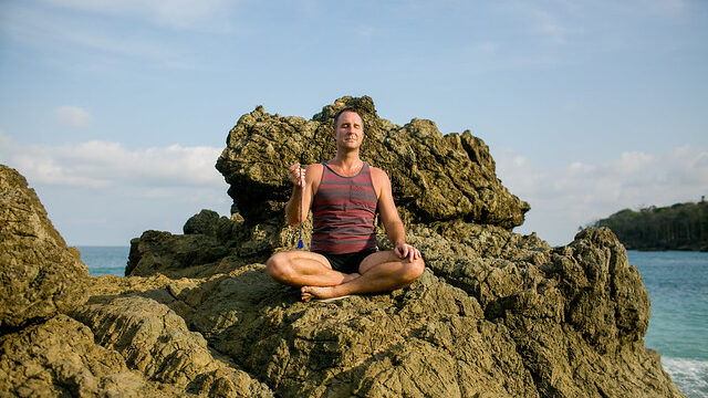 Yogi Aaron meditating with a mala bead necklace on a rock along the ocean in Costa Rica. 