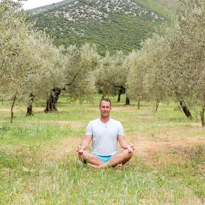 Yogi Aaron sitting in a field surrounded by trees and hills meditating. 