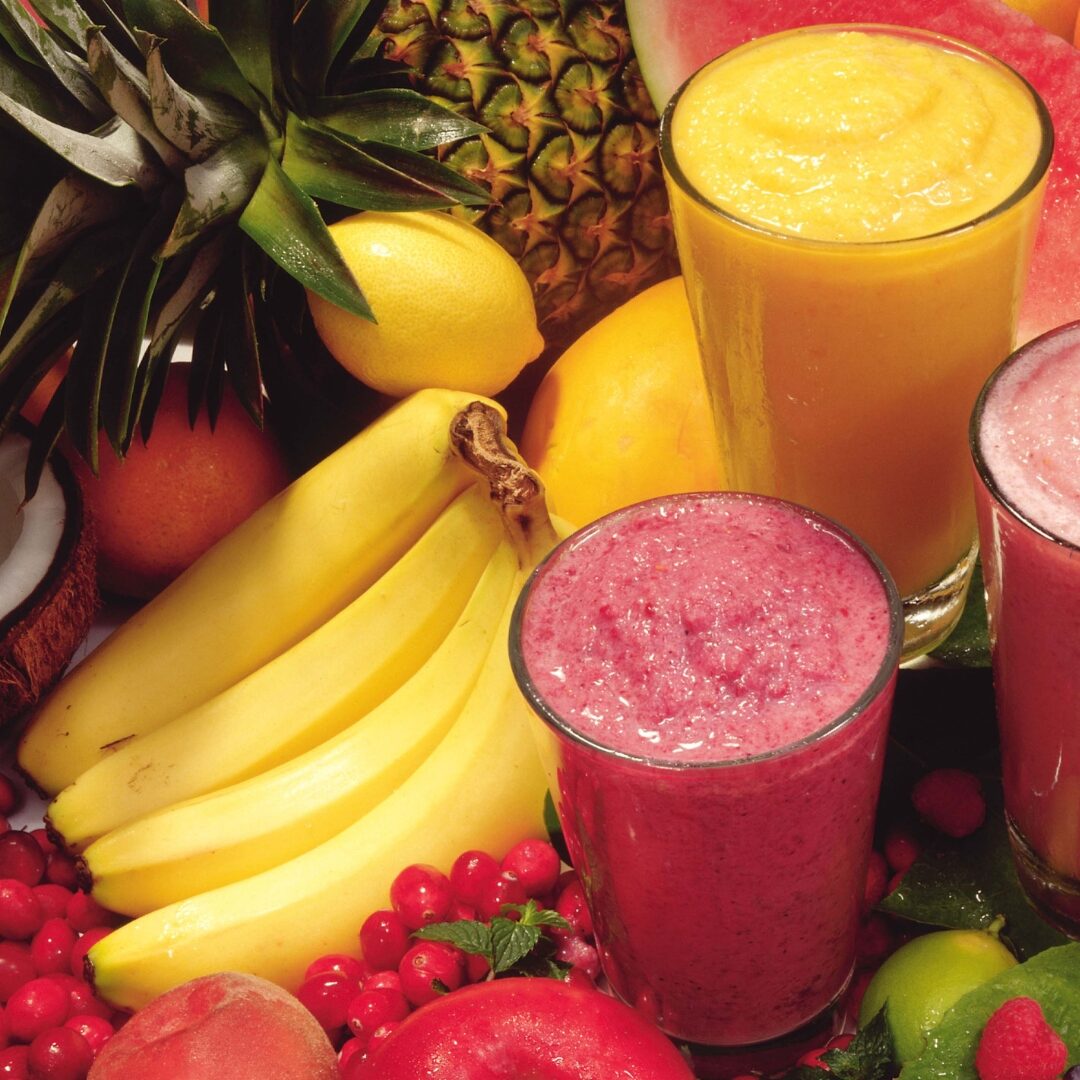 Pink and yellow smoothies surrounded by bananas, pineapples. lemons and raspberries.