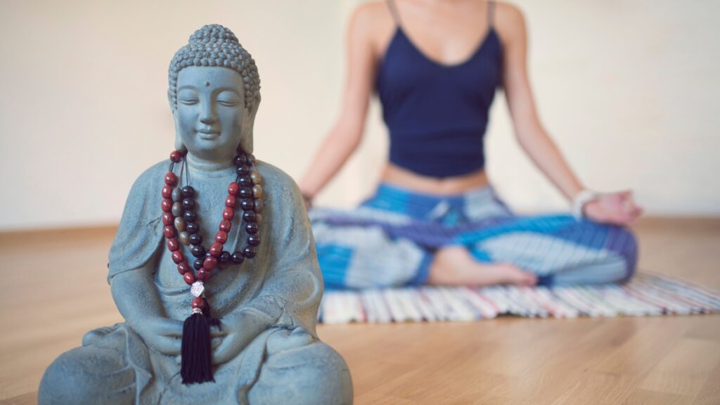 A Buddah statue with mala beads draped around its neck and a woman sitting in a meditative position in the background. 