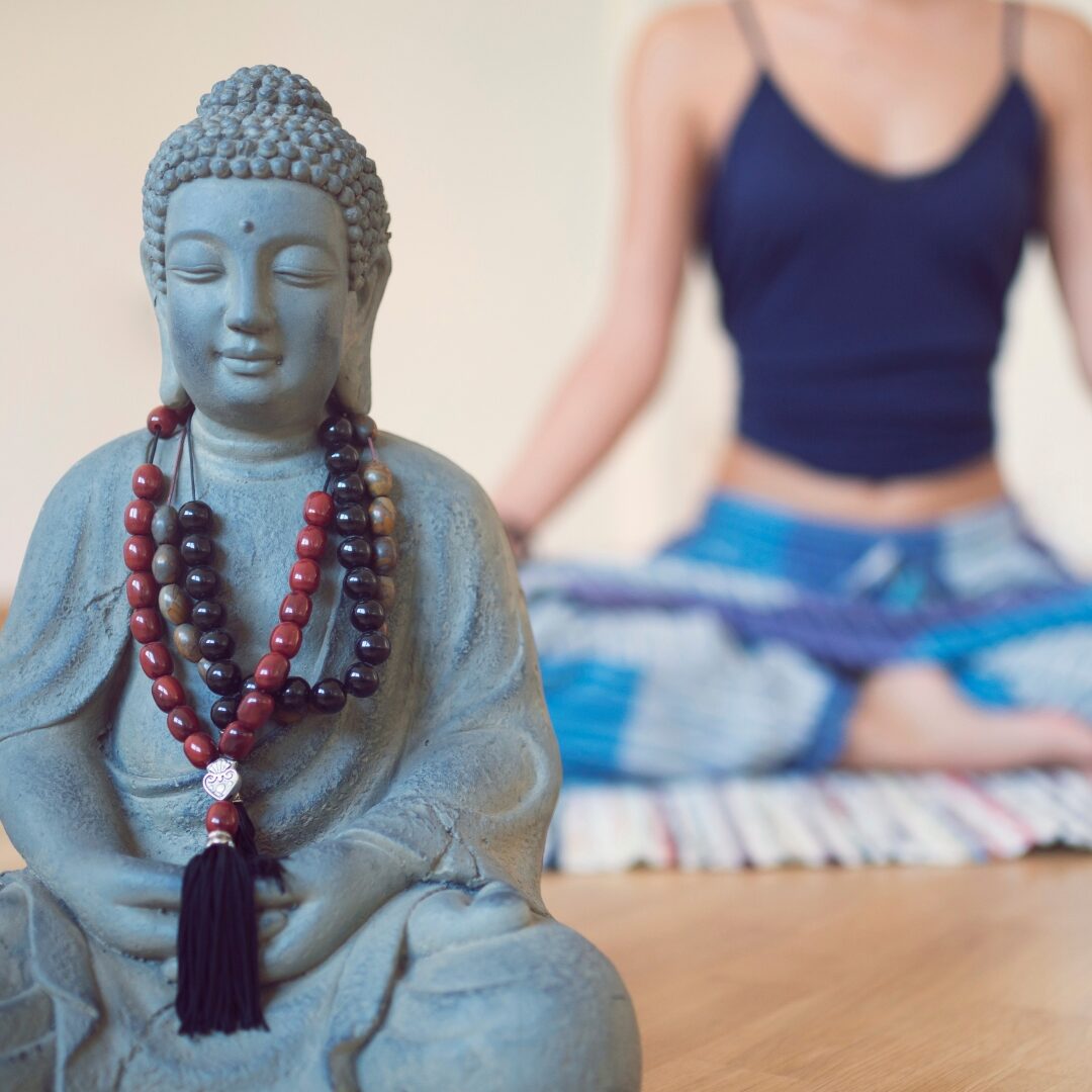 Buddah statue with mala beads draped around its neck and a woman meditating in the background.