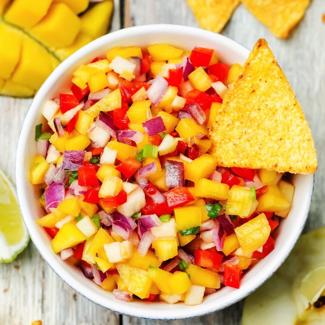 White ceramic bowl of mango salsa with red onions and red peppers garnished with a yellow tortilla chip.