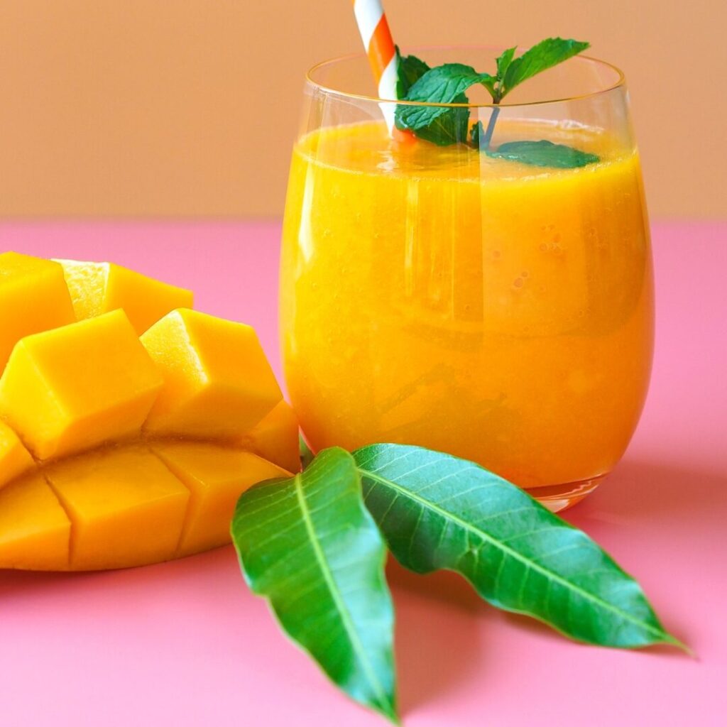 Glass of mango smoothie with a sliced mango and green leaves next to the glass. 