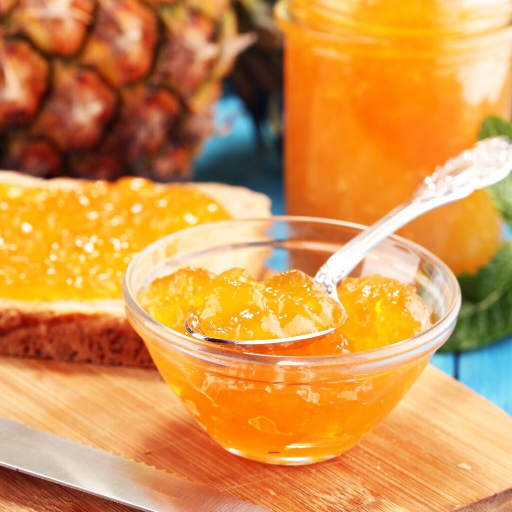 A glass dish of pineapple jam on a cutting board with a slice of bread with jam, a jar of jam and a pineapple in the background.