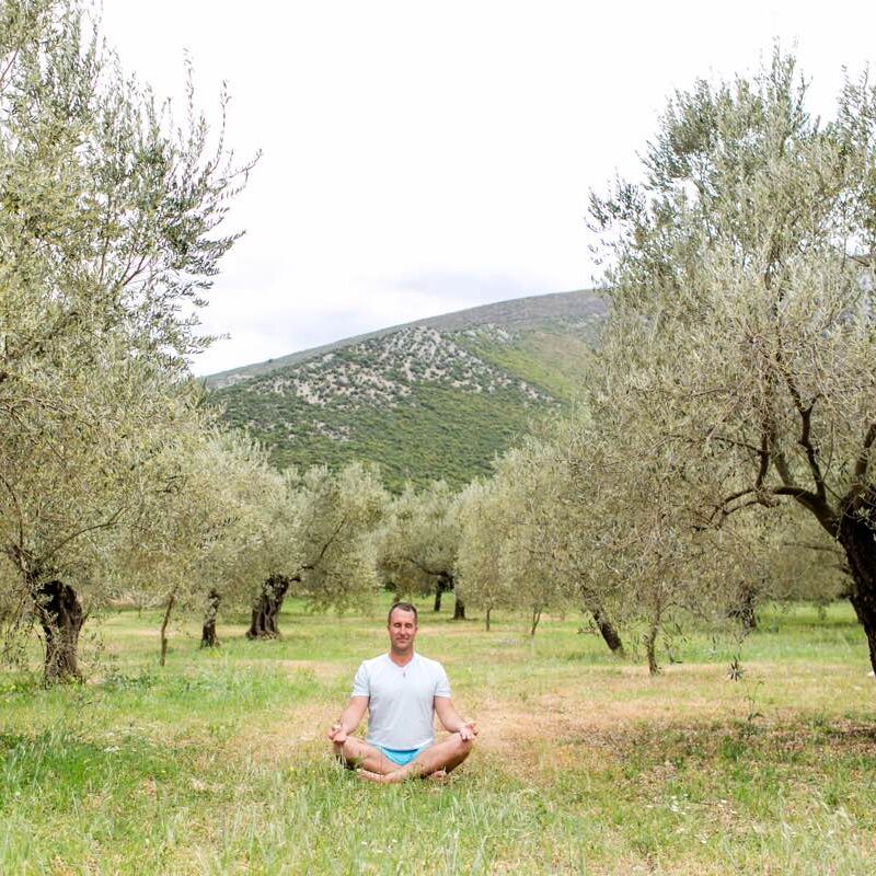 Yogi Aaron sitting in meditation in a field surrounded by trees and hills. 