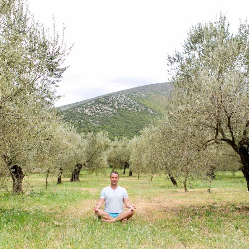 Yogi Aaron sitting in meditation in a field surrounded by trees and hills.