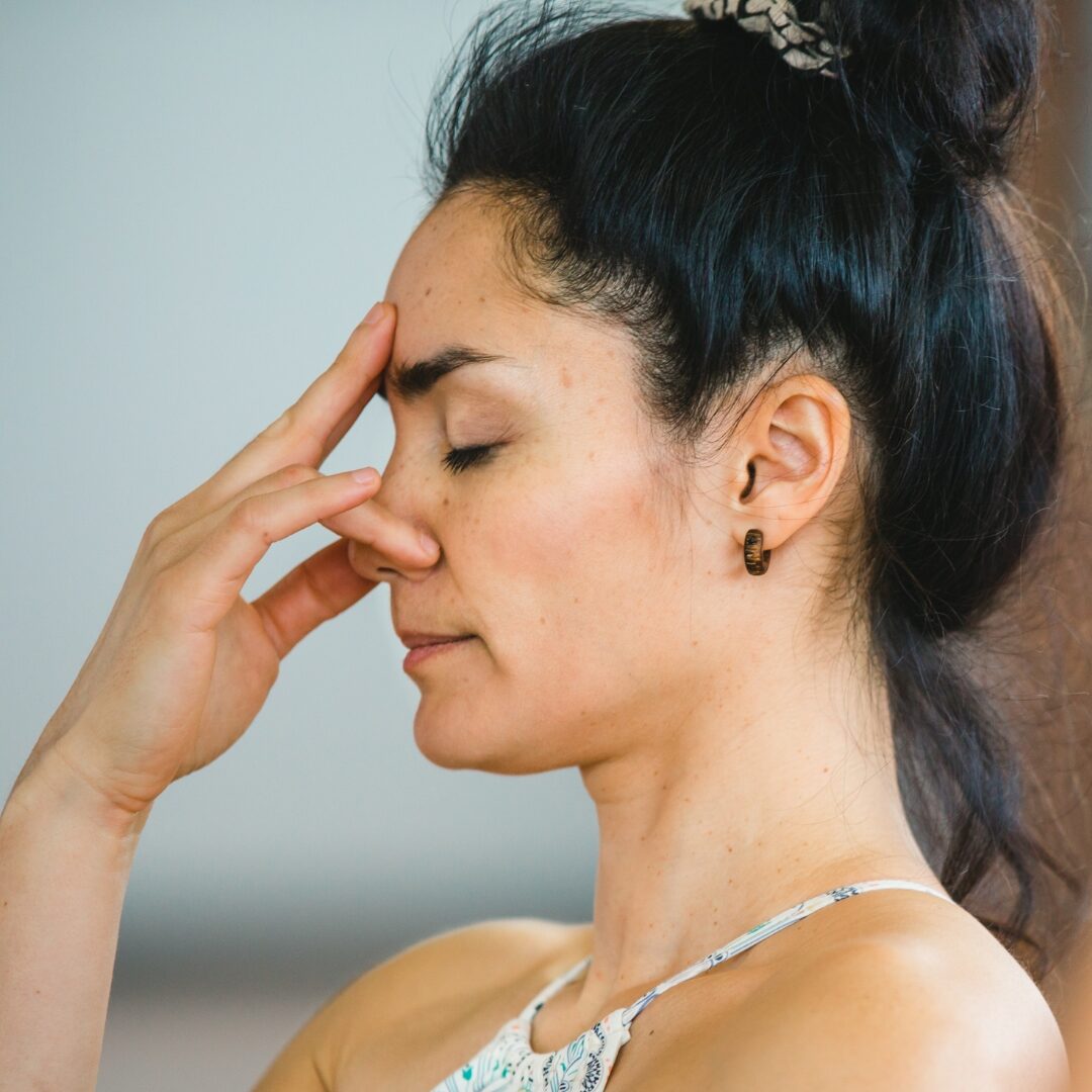 Woman with black hair in a ponytail practicing alternate nostril breathing with her eyes closed. 