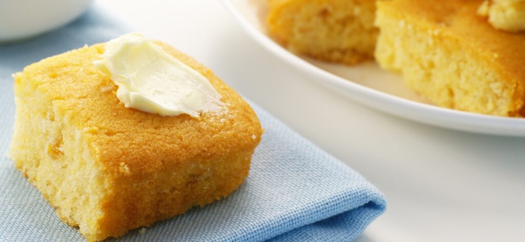 Piece of cornbread with a slab of butter spread on top sitting on a light blue cloth napkin. 