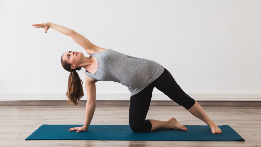 Pregnant woman in black leggings and a gray tank top practicing Gate Pose. 