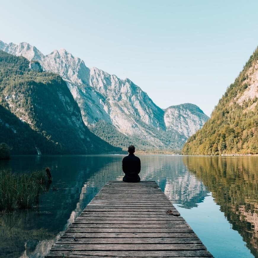 Man sitting on the edge of a dock overlooking a river and mountains. 