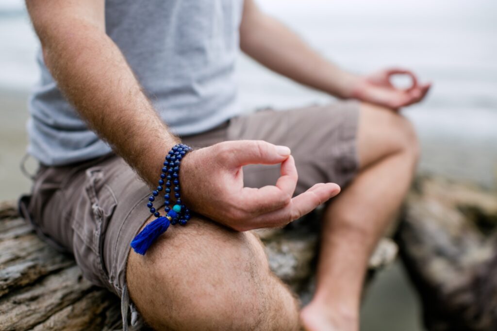 Man sitting in meditation with his hands in a mudra position wearing royal blue mala beads around his right wrist.