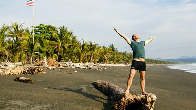 Yogi Aaron standing on a log on the beach with his hands out wide taking in the sun in Costa Rica.