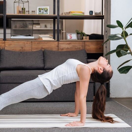 Woman with a long ponytail, white tank top and gray leggings doing a reverse plank in her living room.