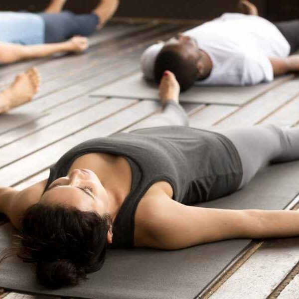Woman with a black tank top and grey pants lying in savasana pose.