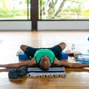 Man with a green tank top and black shorts lying in Savasana pose with his knees in butterfly position and supported by blocks in Blue Osa's yoga shala in Costa Rica. 