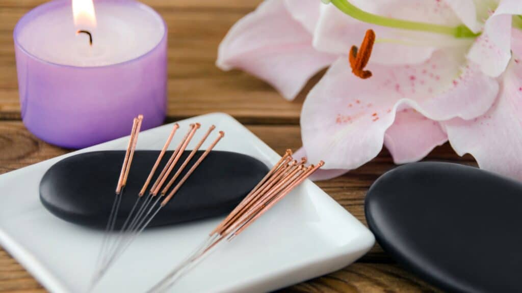 Acupuncture needles sitting on a square white ceramic plate with black massage stones, a lily and a purple flickering candle.